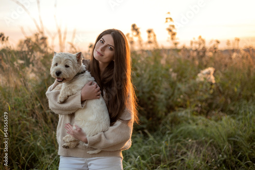 portrait of a beautiful young girl with a dog in her arms, a girl hugs her beloved petam on a walk at the sunset, West Highland White Terrier, copy space