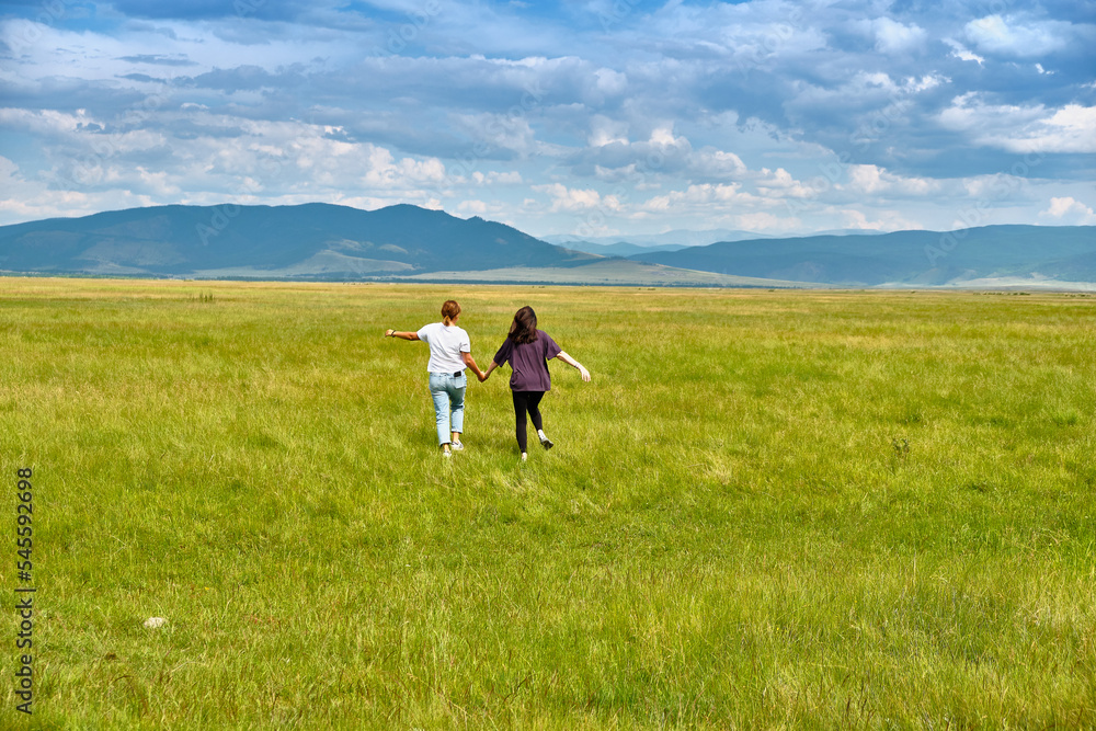 Two girls run through the green grass of the Barguzin Valley in the Republic of Buryatia. View from the back.