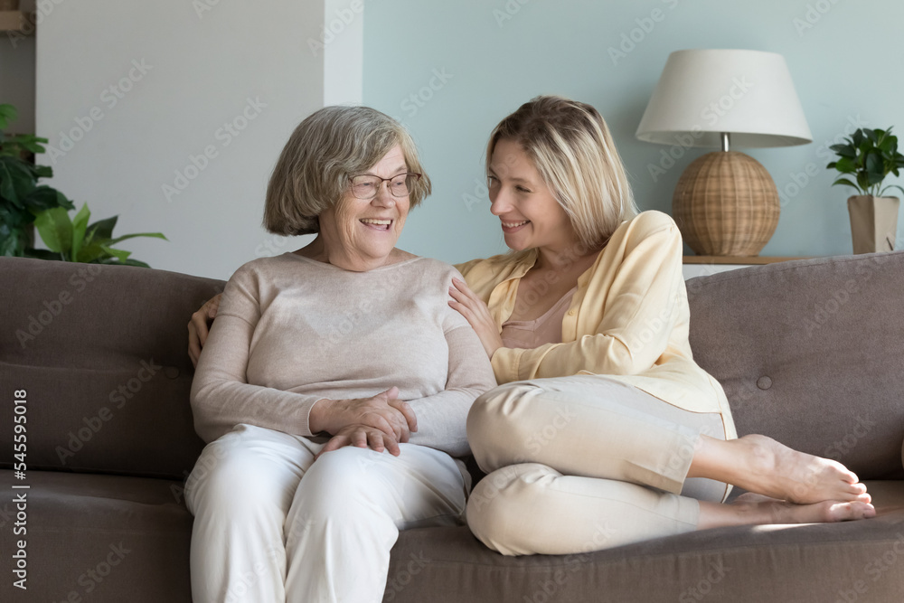 Pretty millennial woman spend time enjoy talk to older grandmother sit on sofa at home. Granny having pleasant conversation with loving grown up granddaughter, share news during her visit. Family ties
