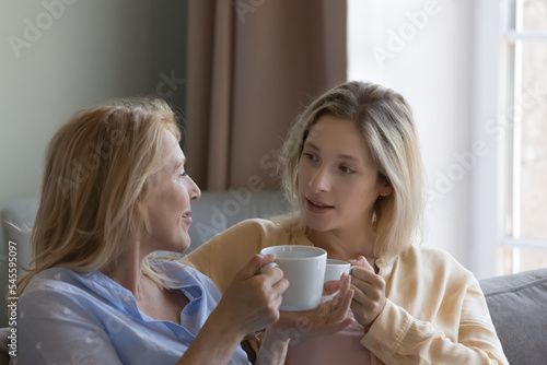 Blonde millennial woman and middle-aged mother holding mugs, drinking coffee, enjoy pleasant friendly conversation, lead trustworthy talk spend leisure together. Understanding, trust, support, family