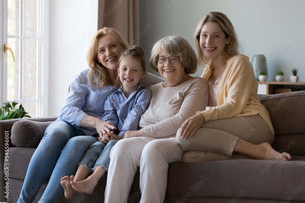 Care, love, multi-generational women family portrait. Senior grandmother, her adult daughter, grown up granddaughter and little cute 6s great-granddaughter smile look at cam relaxing on sofa at home Care, love, multi-generational women family portrait. Senior grandmother, her adult daughter, grown up granddaughter and little cute 6s great-granddaughter smile look at cam relaxing on sofa at home