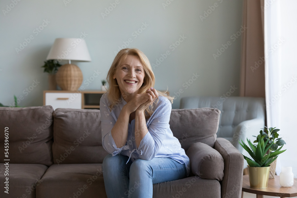 Pretty middle-aged woman posing for picture seated on sofa in modern living room. Portrait of happy, good-looking optimistic female spend carefree leisure at home, enjoy untroubled life on retirement - obrazy, fototapety, plakaty 