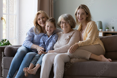 Care, love, multi-generational women family portrait. Senior grandmother, her adult daughter, grown up granddaughter and little cute 6s great-granddaughter smile look at cam relaxing on sofa at home