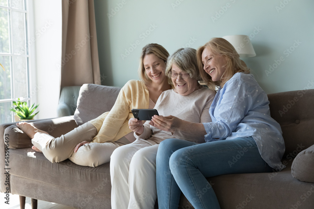 Caring younger generation teach older gen to use modern wireless technology, showing new mobile app for comfort life, spend carefree leisure sit together on sofa at home staring at smartphone screen
