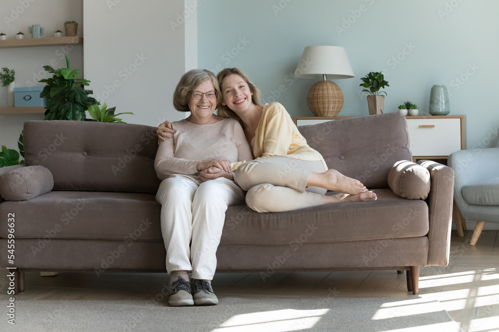 Young adult granddaughter hugs joyful retired older grandmother relax on sofa smile look at camera. Portrait of happy different generations family, showing care, appreciate time together, feeling love