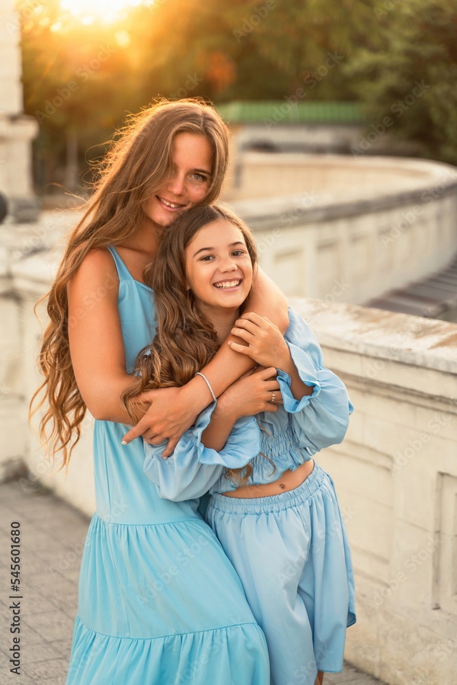 Portrait of mother and daughter in blue dresses with flowing long hair against the backdrop of sunset. The woman hugs and presses the girl to her. They are looking at the camera.