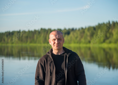 portrait of a confident man in a black hoodie against the background of a river and forest. © Dmitry