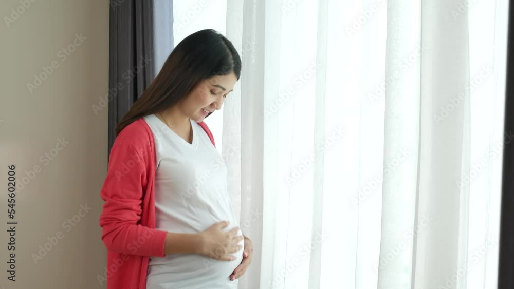 Young Asian pregnant woman standing in bedroom holding belly open white curtain with happy feeling. Young Asian pregnant woman standing in bedroom holding belly open white curtain with happy feeling.