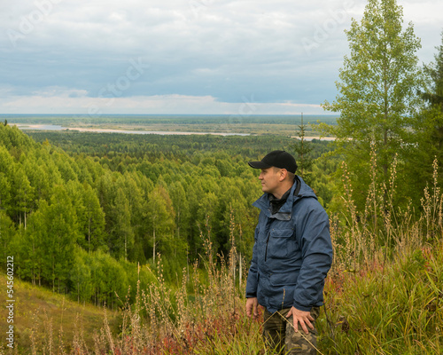 a man in a blue jacket and a black cap on a wooded hill against the background of a forest and a river. © Dmitry