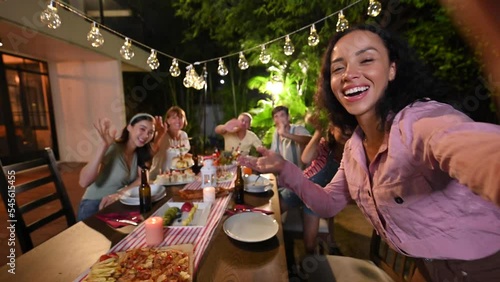 Authentic shot of happy carefree smiling woman making selfie or video call to friends or relatives while at garden party celebration with big family