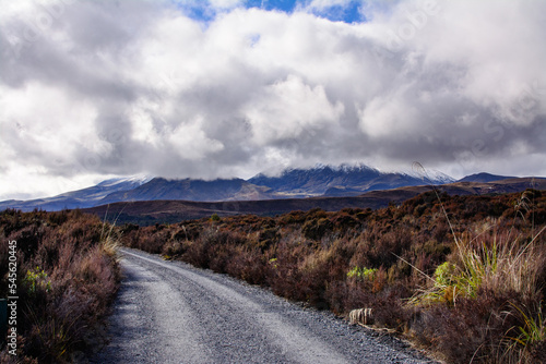 Gravel track curving through desolated high land field in Central Plateau of New Zealand. Snow blanketed volcanic cone of Mount Ngauruhoe covered by low clouds. Tongariro National Park, North Island