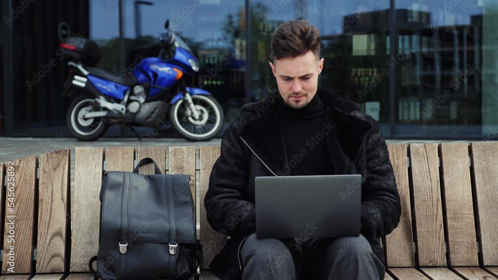 Young man in warm fashion clothes user work on laptop pc computer sit on bench with backpack rest relax on background of blue motorcycle outdoors Mobile office freelance business lifestyle concept. Young man in warm fashion clothes user work on laptop pc computer sit on bench with backpack rest relax on background of blue motorcycle outdoors Mobile office freelance business lifestyle concept.