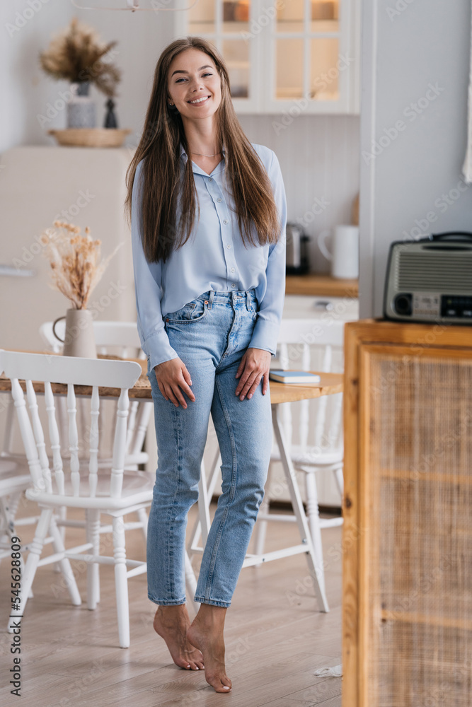 Vertical shoot of gorgeous blonde young woman in casual posing at kitchen smiling wide looks at camera happily stands at kitchen. Beautiful Caucasian fit girl with long loose hair at home. Vertical shoot of gorgeous blonde young woman in casual posing at kitchen smiling wide looks at camera happily stands at kitchen. Beautiful Caucasian fit girl with long loose hair at home.