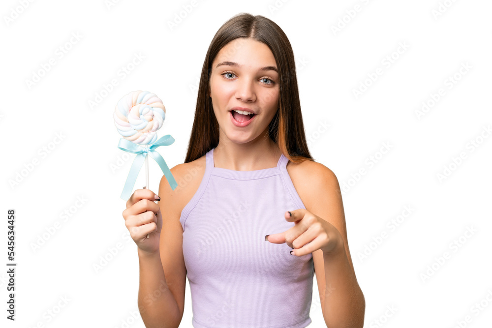 Teenager caucasian girl holding a lollipop over isolated background surprised and pointing front Teenager caucasian girl holding a lollipop over isolated background surprised and pointing front