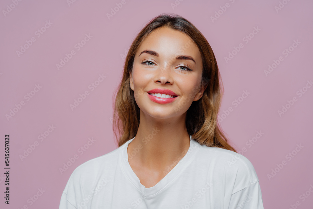 Close up shot of feminine girl smiles gently at camera, wears minimal makeup, has healthy skin, fair hair, feels pleased to get compliment dressed in casual white sweater isolated on purple background Close up shot of feminine girl smiles gently at camera, wears minimal makeup, has healthy skin, fair hair, feels pleased to get compliment dressed in casual white sweater isolated on purple background