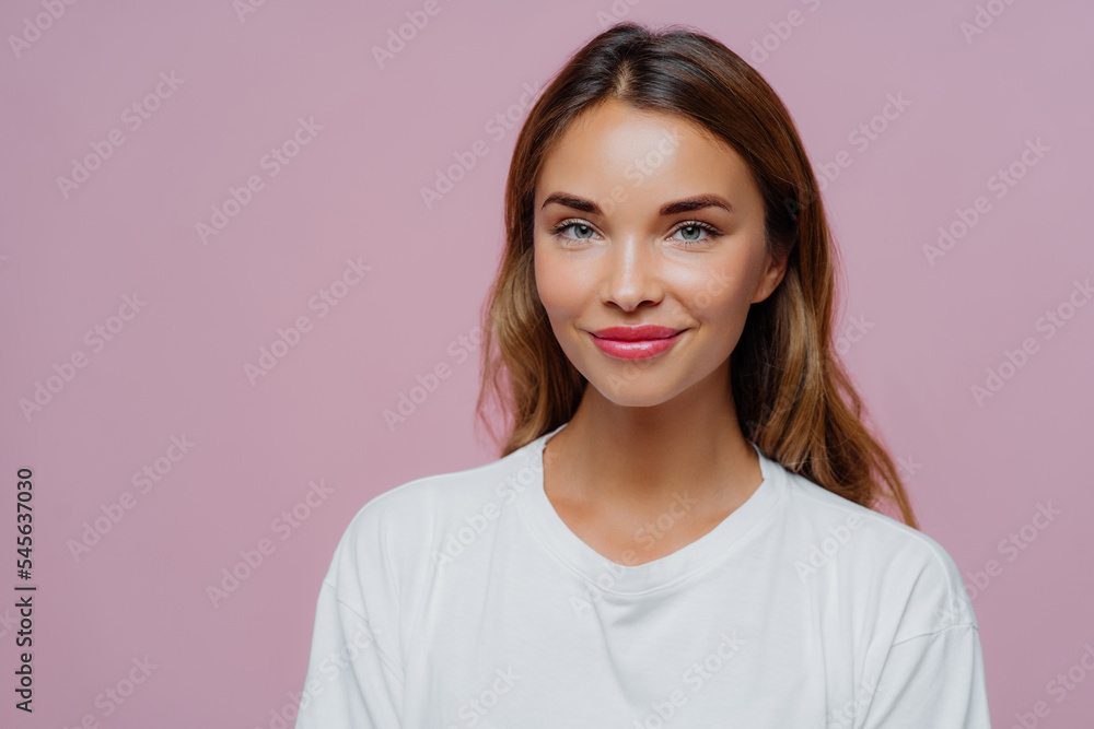 Pretty young woman uses cosmetics for looking young and beautiful, has long hair, looks confidently at camera with happy tender smile, wears casual outfit, poses over purple wall, blank space on right Pretty young woman uses cosmetics for looking young and beautiful, has long hair, looks confidently at camera with happy tender smile, wears casual outfit, poses over purple wall, blank space on right