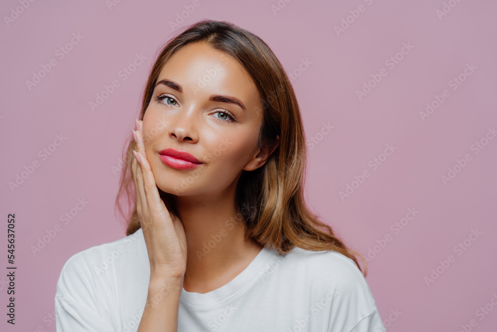 Headshot of lovely female model touches gently cheek, enjoys delicate face skin, wears makeup, has manicure, cares about her beauty, looks straightly at camera, dressed in white jumper, models indoor