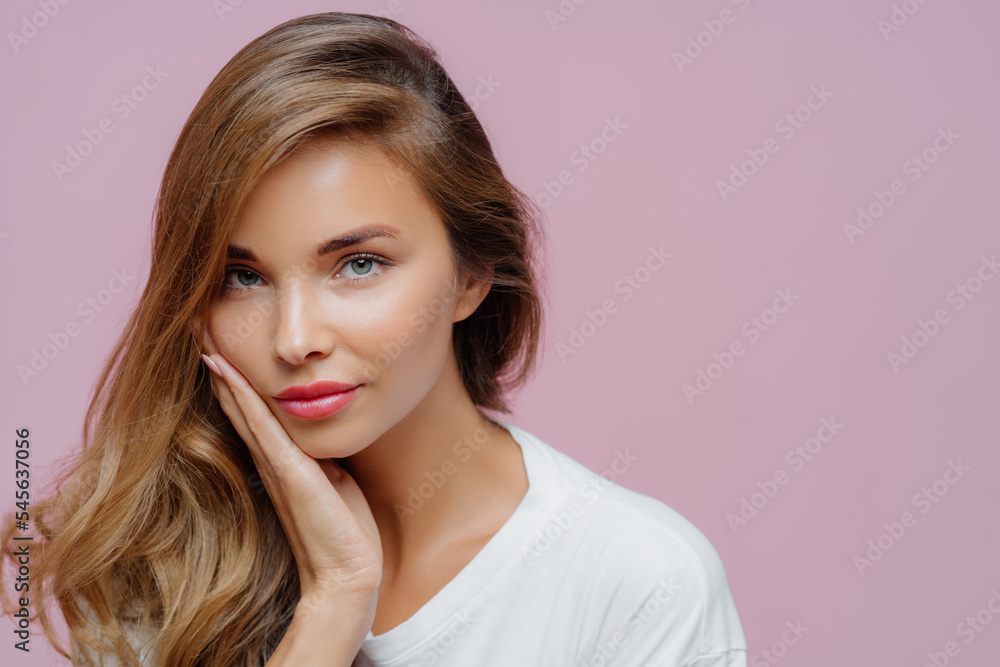 Close up shot of self confident beautiful Caucasian woman has long hair combed on one side, touches cheek with palm, wears minimal professional makeup, looks at herself in mirror, prepares for date Close up shot of self confident beautiful Caucasian woman has long hair combed on one side, touches cheek with palm, wears minimal professional makeup, looks at herself in mirror, prepares for date