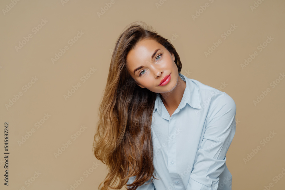 Portrait of pleasant looking female tilts head on right, has long well cared hair, wears blue shirt, has appealing appearance, makeup, isolated over brown background with blank space for information