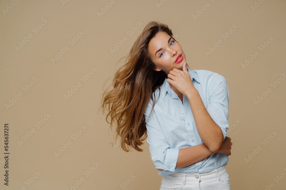 Half length shot of attractive female touches chin, keeps hands partly crossed over chest, tilts head, has long straight hair, dressed in stylish shirt and white jeans, poses against brown background Half length shot of attractive female touches chin, keeps hands partly crossed over chest, tilts head, has long straight hair, dressed in stylish shirt and white jeans, poses against brown background