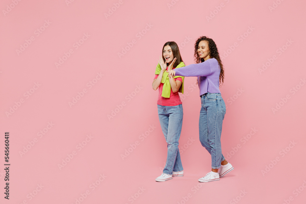 Full body side view young two friends surprised women wear green purple shirts together walk go point finger aside on wokspace area spread hands isolated on pastel plain light pink color background. Full body side view young two friends surprised women wear green purple shirts together walk go point finger aside on wokspace area spread hands isolated on pastel plain light pink color background.