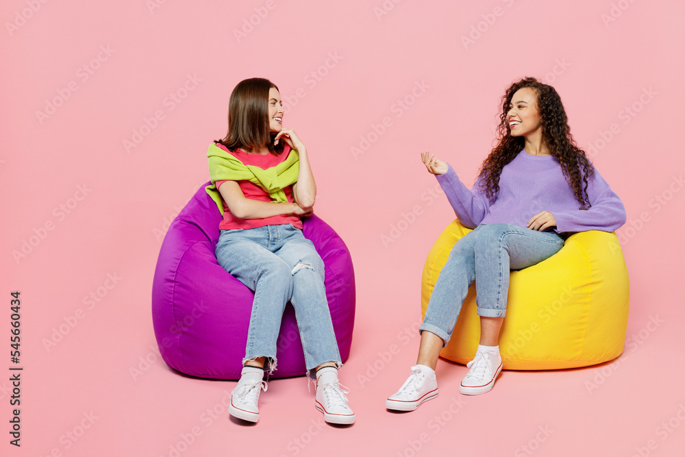 Naklejka premium Full body young two friends positive happy fun cheerful women 20s wears green purple shirts together sit in bag chair talk speak isolated on pastel plain light pink color background studio portrait.