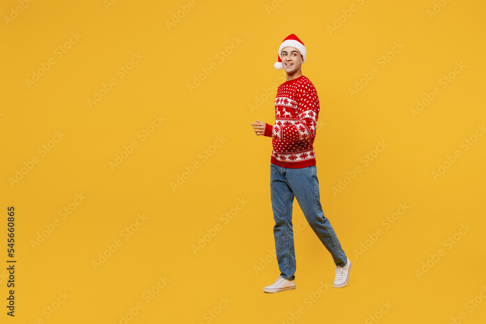 Fototapeta premium Full body side view merry young man wear red knitted Christmas sweater Santa hat posing walk go look aside on area isolated on plain yellow background. Happy New Year 2023 celebration holiday concept.