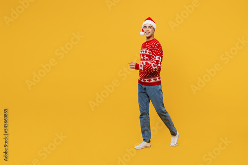 Full body side view merry young man wear red knitted Christmas sweater Santa hat posing walk go look aside on area isolated on plain yellow background. Happy New Year 2023 celebration holiday concept.