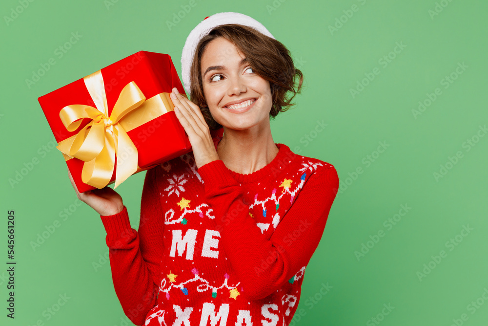 Merry young woman wear xmas sweater Santa hat posing hold red present box with gift ribbon bow look aside on area isolated on plain pastel light green background. Happy New Year 2023 holiday concept. Merry young woman wear xmas sweater Santa hat posing hold red present box with gift ribbon bow look aside on area isolated on plain pastel light green background. Happy New Year 2023 holiday concept.