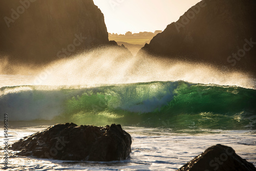 Majestic landscape image of jade turquoise waves crashing onto shore and rocks in Kynance Cove Cornwall with glowing sunrise background and water spray dorplets in wind