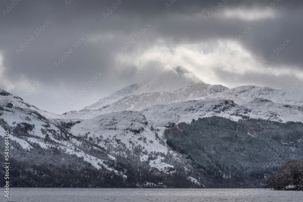 Dramatic Winter landscape image of snow covered trees on shores of Loch Lomond with Ben Lomond mountain looming in background