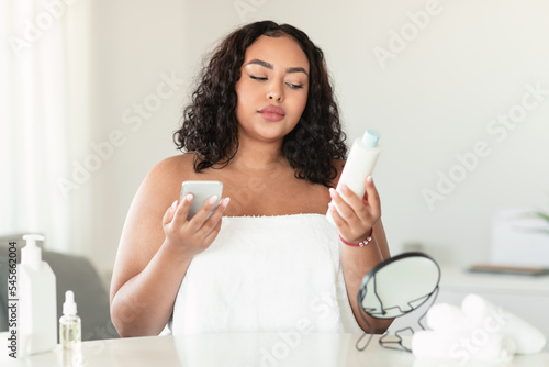 Black body positive lady reading recommendations on lotion bottle and using phone or checking ingredients of cosmetics