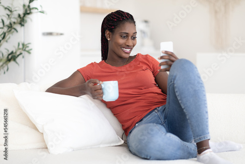 Domestic Relax. Smiling Black Woman Resting On Couch With Smartphone And Coffee