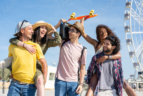 Multiracial group of friends having fun on holiday together, generation z people on vacation listening music and carrying girlfriends on piggyback, travel destination concept