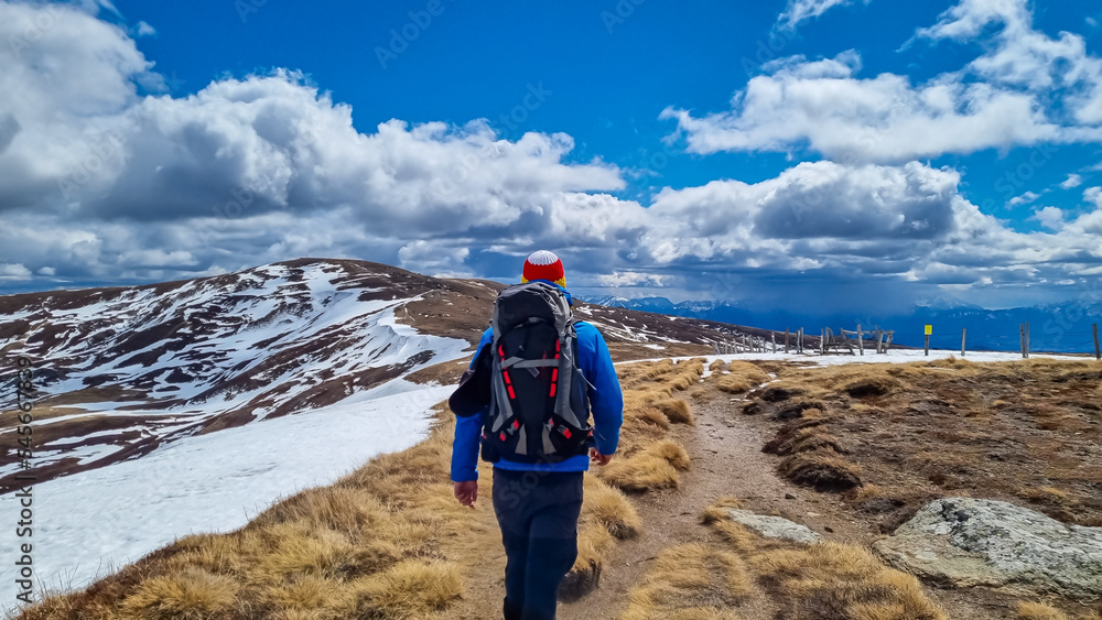 Rear view of man with backpack on hiking trail from Ladinger Spitz to Gertrusk, Saualpe, Lavanttal Alps, Carinthia, Austria, Europe. Trekking on cloudy spring day. Hat in colors of Carinthian flag