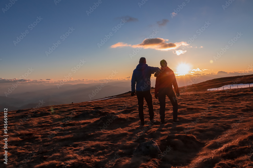Couple standing together on alpine meadow and enjoying beautiful sunset at golden hour on mountain peak Ladinger Spitz, Saualpe, Lavanttal Alps, Carinthia, Austria, Europe. Romantic love atmosphere