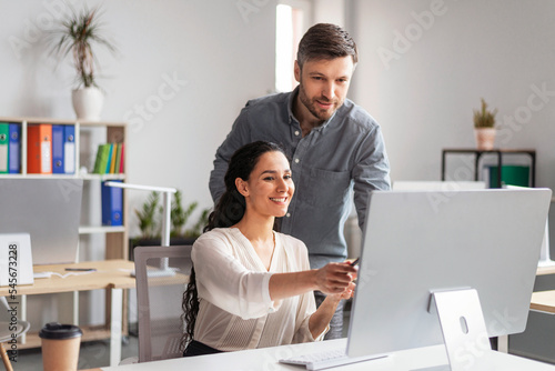 Leinwand Poster Smiling millennial european female showing to man manager with hand in computer