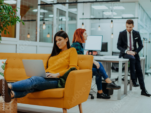 Handsome businesswoman (employee) sitting on a yellow couch while working on her laptop in startup company.