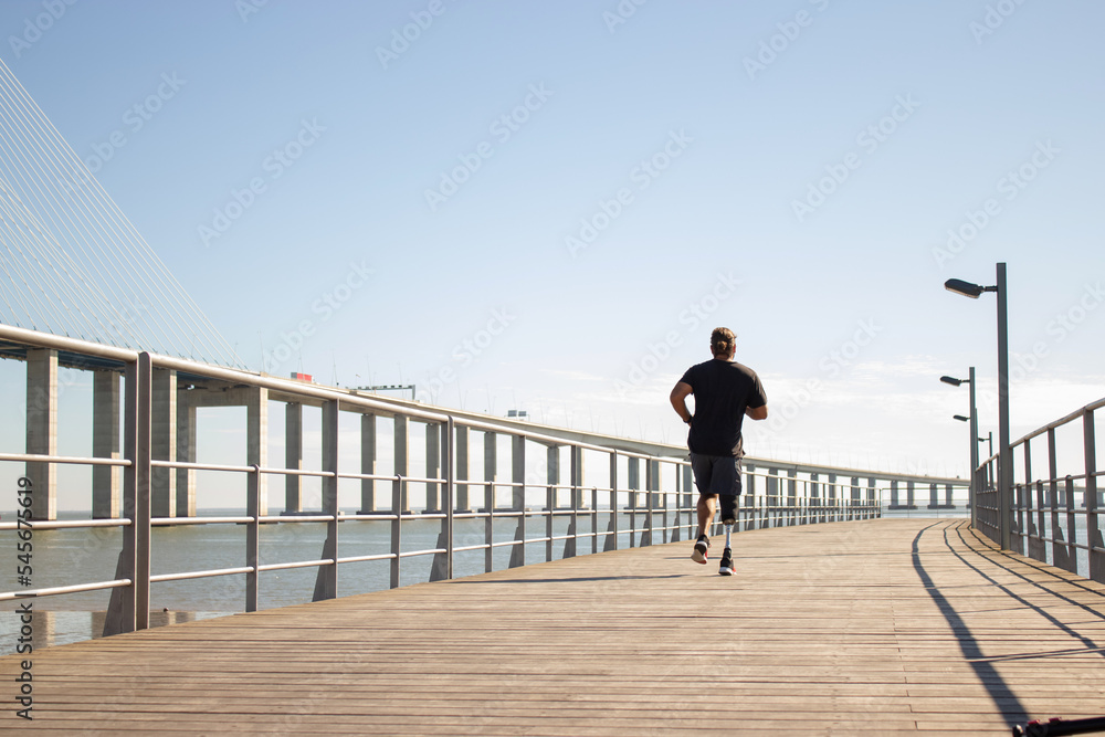 Back view of man with artificial leg jogging on bridge. Sporty man in casual clothes training outdoors in morning running on bridge over sea. Health care, active life of people with disability concept Back view of man with artificial leg jogging on bridge. Sporty man in casual clothes training outdoors in morning running on bridge over sea. Health care, active life of people with disability concept
