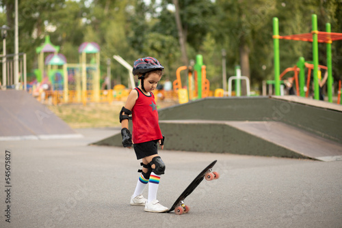 Cute kid girl child with skateboard in a hands standing in a skatepark and smiling. Child performs tricks. Summer sport activity concept.