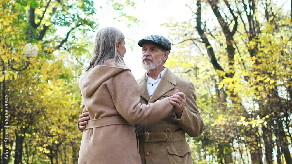 Lovely couple of elderly elegant man and beautiful loving woman slowly dancing in park in sunny autumn day. Happy wealthy husband and wife wearing coats spending time together on fresh air. Lovely couple of elderly elegant man and beautiful loving woman slowly dancing in park in sunny autumn day. Happy wealthy husband and wife wearing coats spending time together on fresh air.