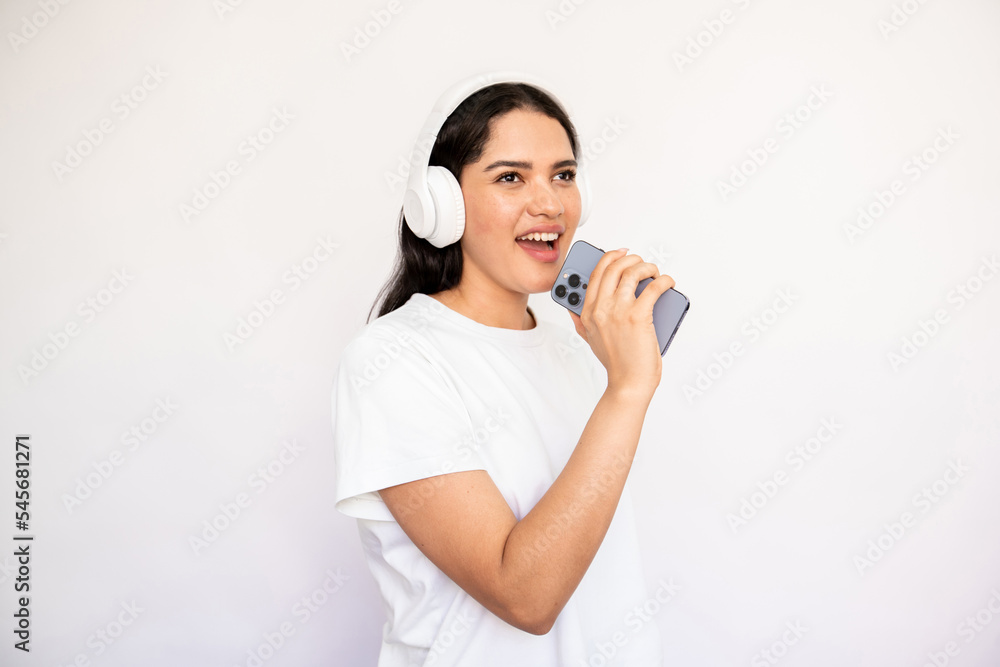 Portrait of happy young woman in headphones recording message with mobile phone. Caucasian lady wearing white T-shirt listening to music and singing. Mobile technology and leisure concept Portrait of happy young woman in headphones recording message with mobile phone. Caucasian lady wearing white T-shirt listening to music and singing. Mobile technology and leisure concept