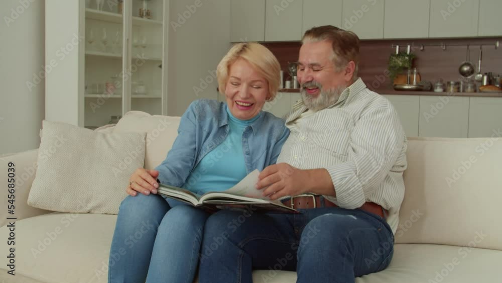 Joyful attractive senior woman and cheerful handsome mature man sitting on couch, watching family photo album, sharing and remembering happy and funny moments while retired couple relaxing at home. Joyful attractive senior woman and cheerful handsome mature man sitting on couch, watching family photo album, sharing and remembering happy and funny moments while retired couple relaxing at home.