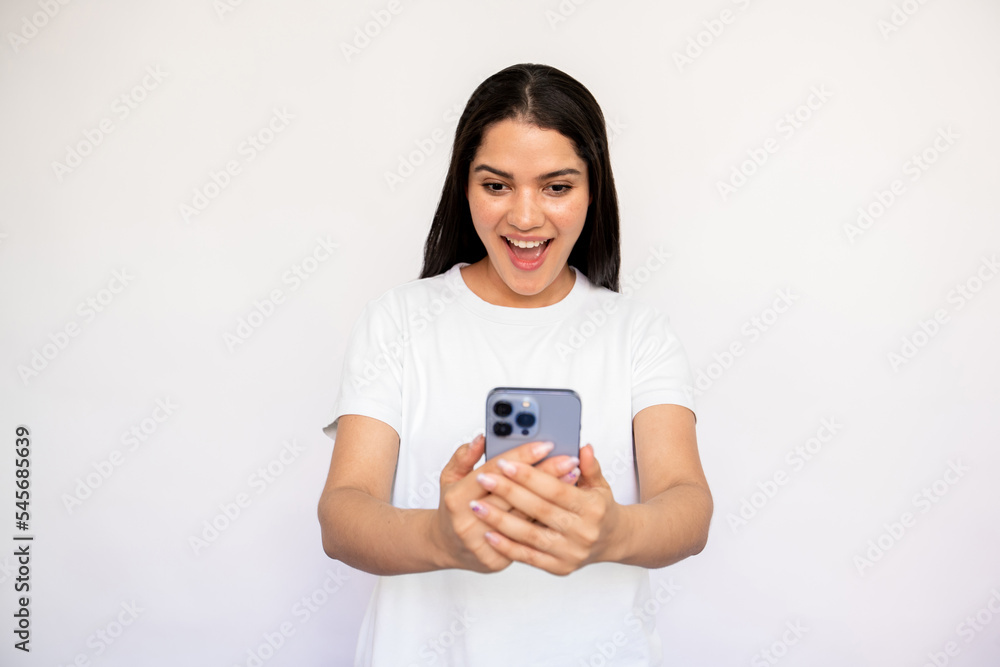 Portrait of excited young woman reading newsletter on smartphone over white background. Caucasian lady wearing white T-shirt surfing internet on mobile phone. Mobile technology concept Portrait of excited young woman reading newsletter on smartphone over white background. Caucasian lady wearing white T-shirt surfing internet on mobile phone. Mobile technology concept