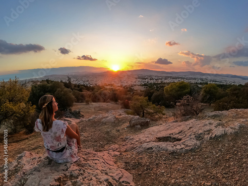 Tourist woman watching sunset over city of Athens seen from Filopappou Hill (hill of muses), Athens, Attica, Greece, Europe. Athens cityscape and Aegean sea. Beautiful sunset point with aerial view