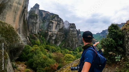 Active man with backpack standing under massive rock formation pinnacles near village Kastraki on mystical hiking trail in Kalambaka, Meteora, Thessaly district, Greece, Europe. Rock climbing activity