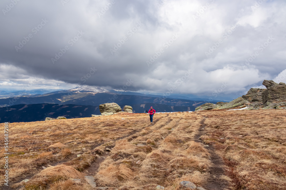 Front view of woman with hiking backpack on alpine meadow with rock formations near Ladinger Spitze, Saualpe, Lavanttal Alps, Carinthia, Austria Europe. Trekking on cloudy early spring day. Wanderlust
