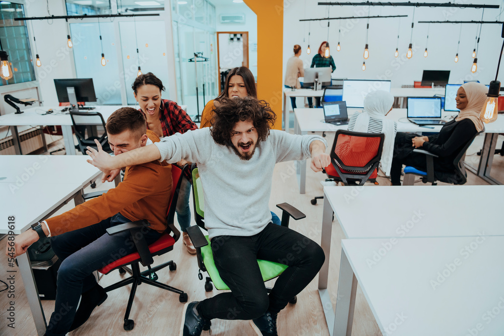 Fototapeta premium Team building and office fun. Young cheerful businesspeople in smart casual wear having fun while racing on office chairs and smiling.