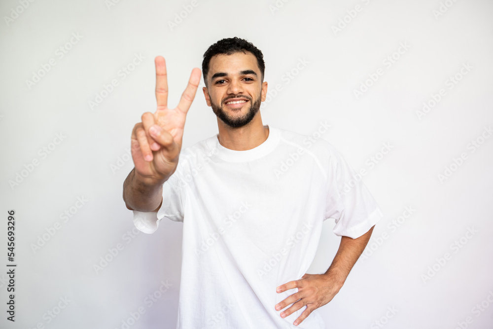 Portrait of happy young man with victory gesture. Pleased Caucasian male model with dark hair and beard in white T-shirt looking at camera, smiling and doing peace sign. Success, optimism concept Portrait of happy young man with victory gesture. Pleased Caucasian male model with dark hair and beard in white T-shirt looking at camera, smiling and doing peace sign. Success, optimism concept