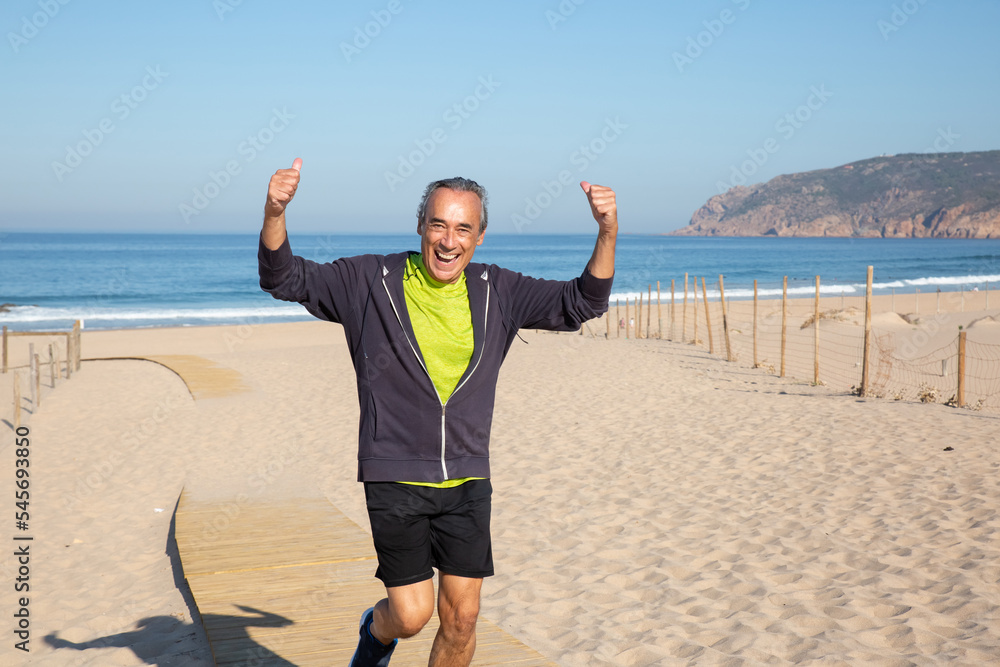 Portrait of sporty aged man spending time on sea beach. Smiling man in sportswear demonstrating his strength and health for camera after active training. Happy life in motion for aged people concept Portrait of sporty aged man spending time on sea beach. Smiling man in sportswear demonstrating his strength and health for camera after active training. Happy life in motion for aged people concept
